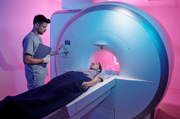 Молодой врач в форме нажимает кнопку запуска МРТ машины с пациентом - Фото, изображение