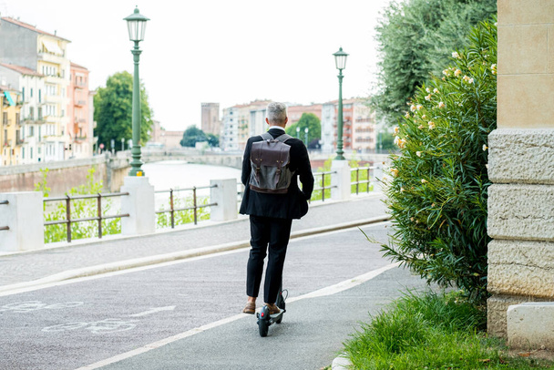 Şehirde işe giderken elektrikli scooter kullanan takım elbiseli genç bir işadamı. Ekolojik ulaşım kavramı - Fotoğraf, Görsel