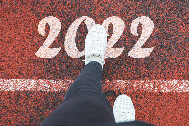 Feliz Año Nuevo 2022. Las piernas de las mujeres cruzan la línea blanca y entran en 2022. Vista superior de las zapatillas blancas en la cinta de correr con texto 2022, concepto de celebración de año nuevo - Foto, Imagen