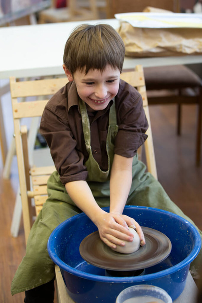 Ένα μικρό αγόρι φτιάχνει ένα πήλινο προϊόν στον τροχό ενός αγγειοπλάστη.. - Φωτογραφία, εικόνα
