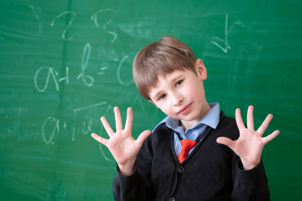 Estudiante infantil en el aula. Feliz colegial en la pizarra verde muestra su grado diez con las manos. Educación primaria. - Foto, imagen