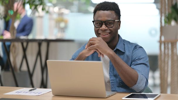 Hombre africano sonriendo en la cámara mientras trabaja en el ordenador portátil - Foto, imagen