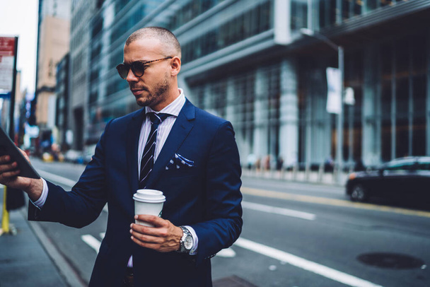 Formally dressed bald businessman standing on sidewalk and stretching out hand with tablet while reading information and holding cup of coffee - Foto, Imagem
