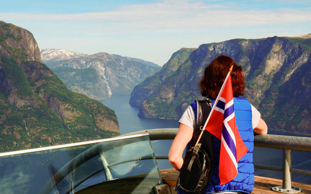 Female tourist with norwegian flag enjoying scenic view over fjord Aurlandsfjord from Stegastein viewing platform. National tourist route Aurlandsfjellet. - Фото, зображення