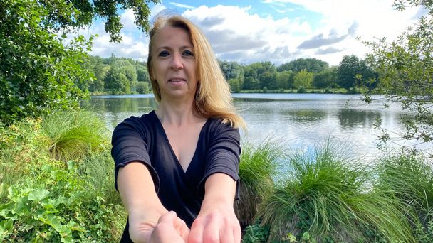 Eine blonde Frau hält jemandem die Hand. Glückliche weibliche Person, die Kamera nach draußen schaut. Fluss oder See im Hintergrund. - Foto, Bild