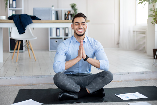 Gestión del estrés en el trabajo. Empleado árabe pacífico meditando, haciendo gesto namaste en esterilla de yoga en la oficina - Foto, Imagen