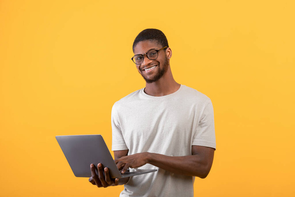 Excited black freelancer man using laptop, standing in studio over yellow background and smiling to camera, copy space - Фото, изображение