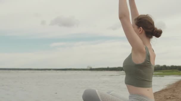 Langzaam schot van jonge ontspannen vrouw in sportkleding zittend in lotus poseren door water op zandstrand op winderige ochtend alleen, beoefenen van yoga en genieten van frisse lucht - Video