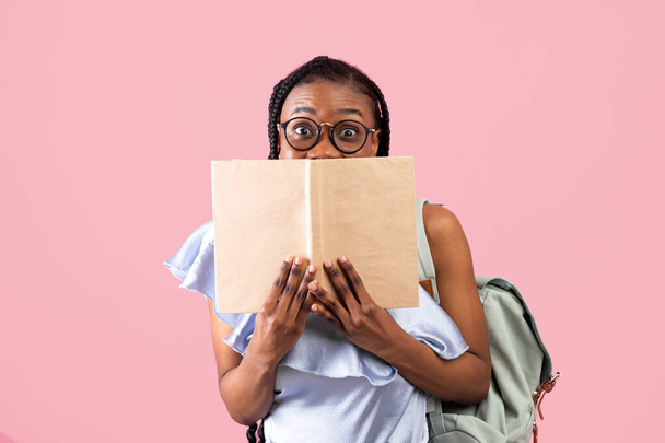 Schockierte afroamerikanische Studentin mit Rucksack versteckt sich hinter aufgeschlagenem Buch auf rosa Studiohintergrund - Foto, Bild