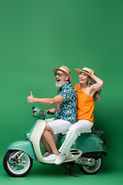 happy middle aged man showing thumb up while riding with asian wife on moped on green  - Fotoğraf, Görsel
