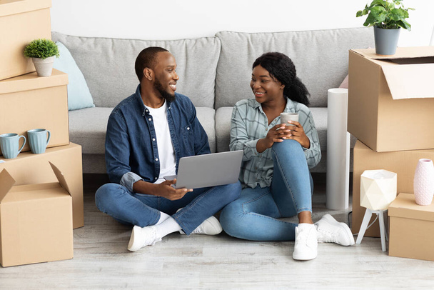 Couple noir joyeux relaxant avec ordinateur portable après avoir déménagé dans un nouvel appartement - Photo, image