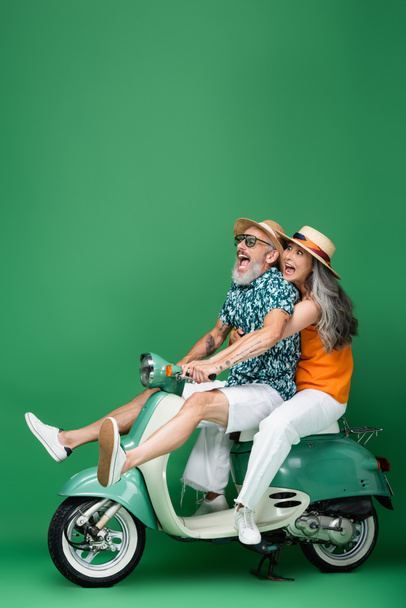 scared and multiethnic middle aged couple in sun hats riding moped on green - Фото, зображення