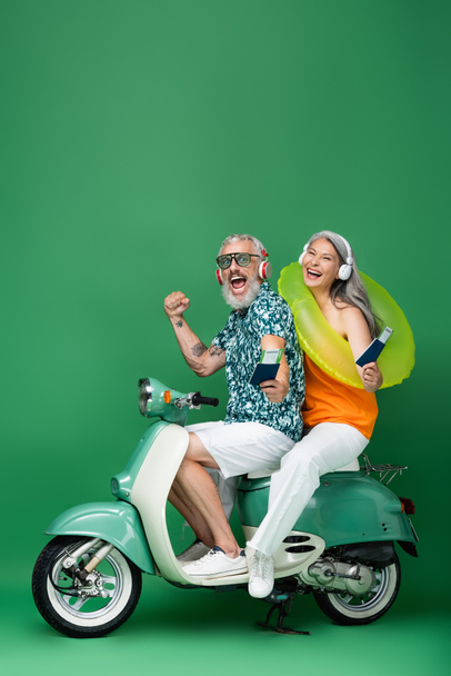 multiethnic and middle aged couple in wireless headphones holding passports with air tickets and riding moped on green - Photo, Image