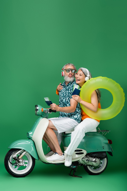 multiethnic middle aged couple in wireless headphones holding passports with air tickets and riding motor scooter on green - Photo, Image