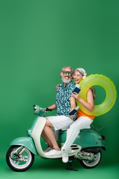 happy and multiethnic mature couple in wireless headphones holding passports with air tickets and riding motor scooter on green - Photo, Image