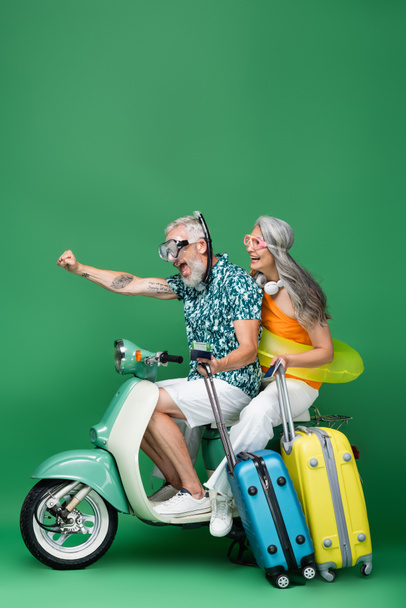 ámulatba ejtő és többnemzetiségű középkorú házaspár, akik zöld mopeden utazva útlevéllel rendelkeznek repülőjegyekkel és csomagokkal - Fotó, kép