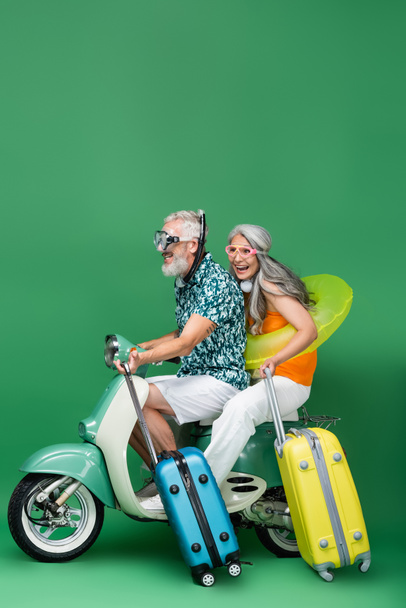 multiethnisches und fröhliches Paar mittleren Alters in Schutzbrille, das Gepäck hält, während es Motorroller auf Grün fährt - Foto, Bild