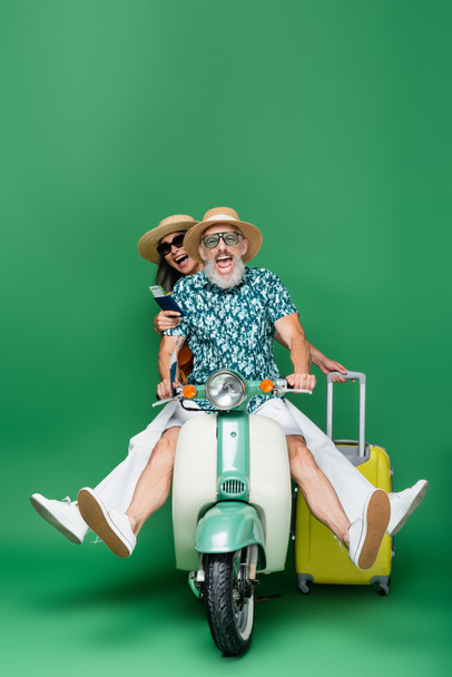 pareja interracial y positiva de mediana edad en sombreros con pasaportes con billetes de avión mientras monta ciclomotor en verde - Foto, Imagen