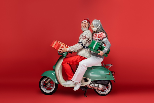 Χαρούμενη μεσήλικη και Ασιάτισσα με ωτοασπίδες που κρατάει χριστουγεννιάτικα δώρα ενώ καβαλάει μοτοποδήλατο με τον άντρα της στο κόκκινο  - Φωτογραφία, εικόνα