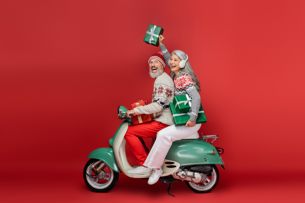 cheerful middle aged asian woman in ear muffs holding christmas presents while riding motor scooter with excited husband on red  - Foto, afbeelding