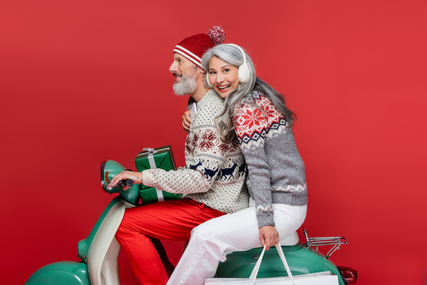 happy and interracial mature couple with christmas presents riding moped on red - Photo, Image