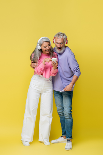 full length of happy interracial and middle aged woman in wireless headphones pointing at smartphone near surprised husband on yellow - Photo, Image