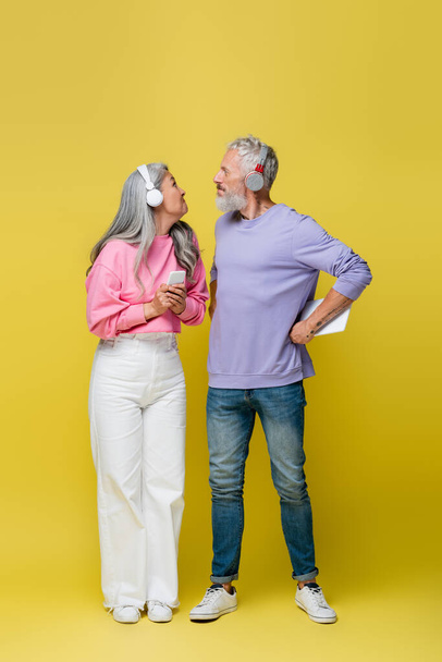 full length of funny interracial and middle aged couple in wireless headphones holding gadgets and looking at each other on yellow  - Fotoğraf, Görsel