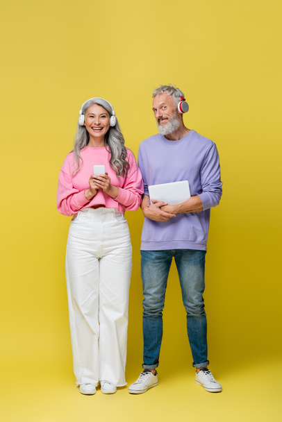 full length of multiethnic and smiling middle aged couple in wireless headphones holding gadgets on yellow  - Fotoğraf, Görsel