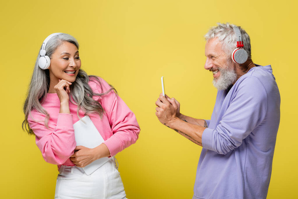 onnellinen keski-ikäinen ja monikansallinen mies langattomissa kuulokkeissa ottaen kuva iloinen vaimo eristetty keltainen - Valokuva, kuva