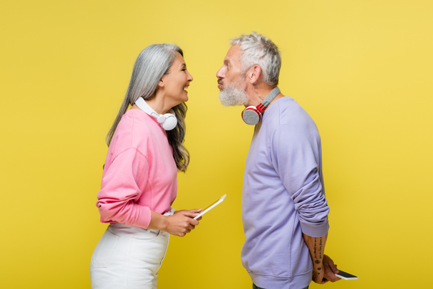 side view of funny multiethnic and middle aged couple in wireless headphones looking at each other and grimacing isolated on yellow  - Фото, изображение
