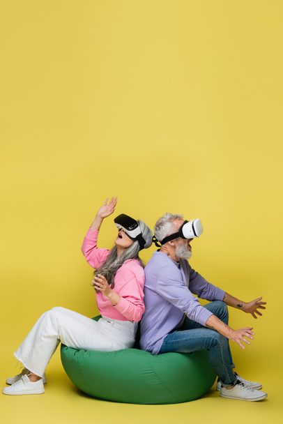 interracial and surprised middle aged couple in vr headsets sitting in bean bag chair while gaming on yellow  - Φωτογραφία, εικόνα