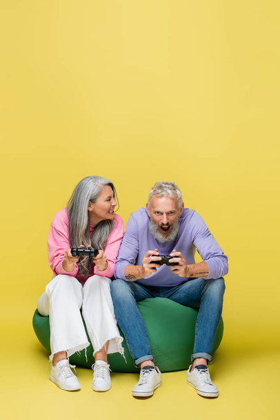 KYIV, UCRANIA - 10 de agosto de 2021: mujer interracial y de mediana edad sonriendo los dientes mientras juega videojuegos con su esposo sorprendido en amarillo  - Foto, Imagen