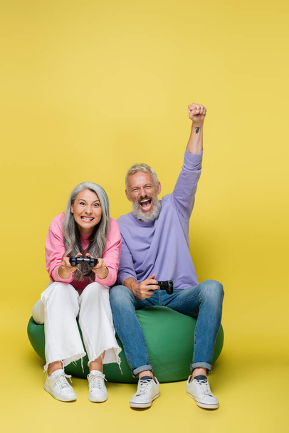 KIEW, UKRAINE - 10. AUGUST 2021: Verblüfftes Paar mittleren Alters hält Steuerknüppel in der Hand und spielt Videospiel auf Bean Bag Stuhl auf gelb  - Foto, Bild