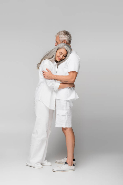 full length of happy interracial and mature couple hugging on grey - Fotó, kép
