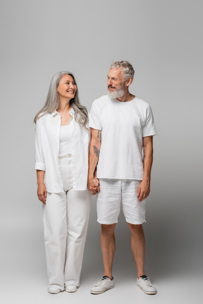full length of happy interracial and mature couple holding hands on grey - Photo, Image