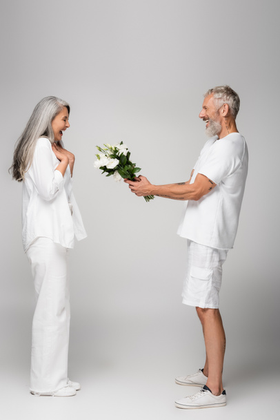 side view of happy middle aged man giving bouquet of flowers to amazed asian wife on grey - Foto, imagen