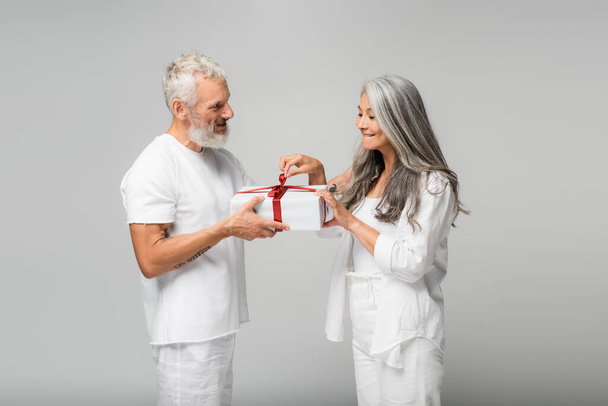 happy middle aged man holding wrapped gift box near curious asian wife isolated on grey - Photo, image