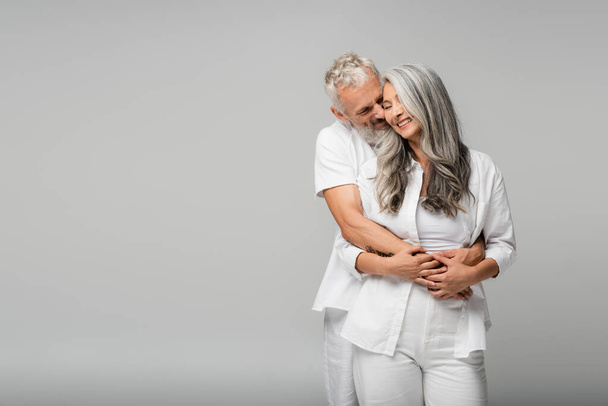 cheerful mature man with closed eyes hugging pleased asian wife isolated on grey - Photo, Image