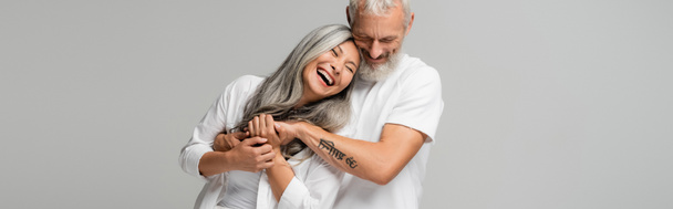 счастливый зрелый мужчина с закрытыми глазами обнимает радостную азиатскую жену изолированную на сером, баннер - Фото, изображение