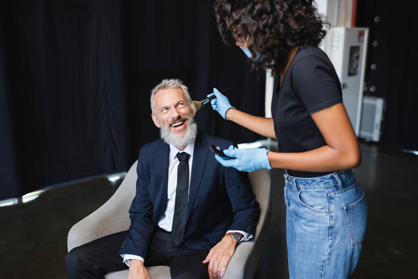 african american makeup artist in medical mask and latex gloves applying face powder on cheerful businessman  - Foto, imagen