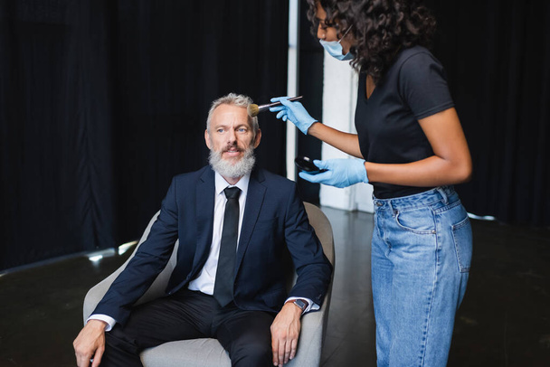 curly african american makeup artist in medical mask and latex gloves applying face powder on businessman  - Photo, Image