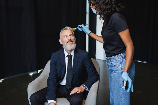 curly african american makeup artist in medical mask and latex gloves applying face powder on smiling businessman  - Photo, Image