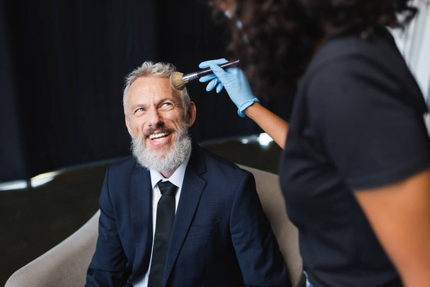 blurred african american makeup artist in latex glove applying face powder on happy businessman  - Photo, Image