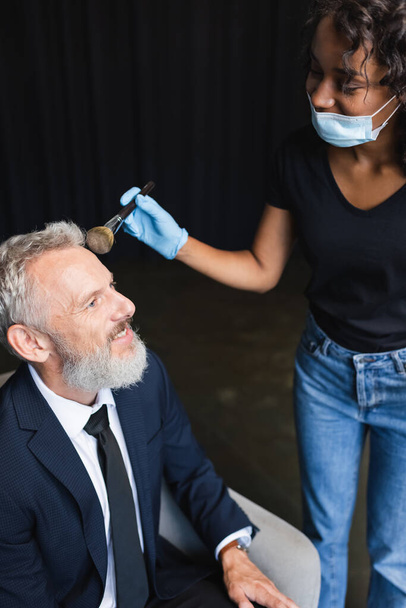 curly african american makeup artist in medical mask and latex glove applying face powder on smiling businessman  - Foto, Bild
