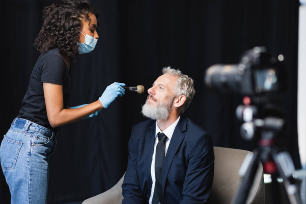 african american makeup artist in medical mask and latex gloves applying face powder on businessman near blurred digital camera - Zdjęcie, obraz