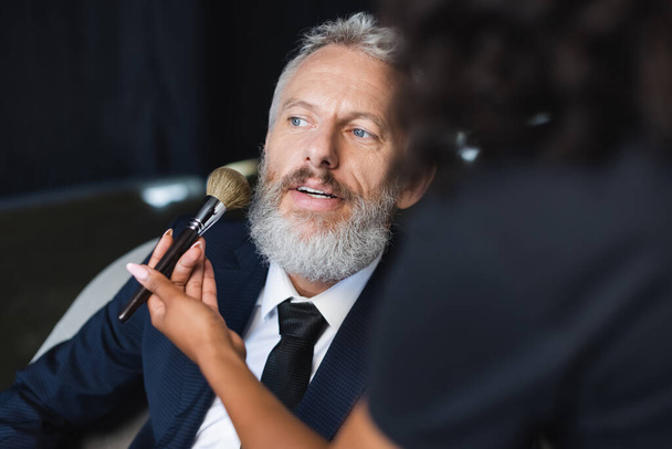 blurred african american makeup artist applying face powder on businessman in interview studio - Photo, Image