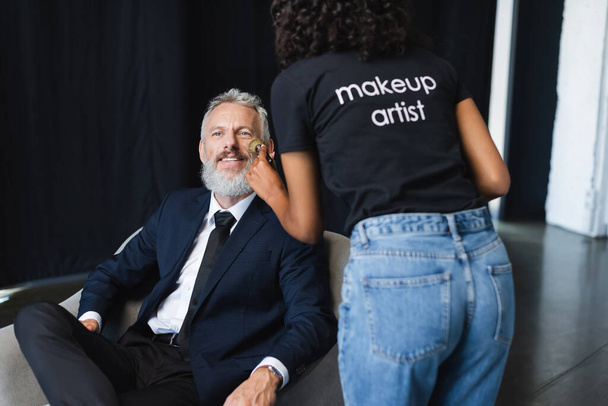 curly african american makeup artist in t-shirt with lettering applying face powder on happy businessman in interview studio - Photo, Image