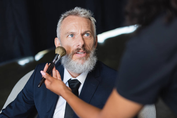 blurred african american makeup artist applying face powder on businessman in formal wear - Photo, Image