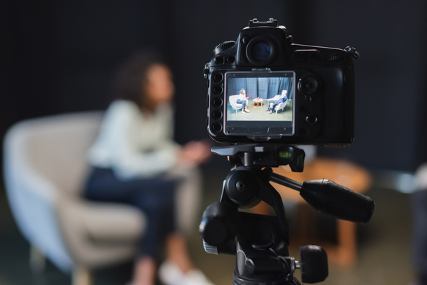 Digitalkamera mit schwarzgelocktem afrikanisch-amerikanischem Journalisten im Gespräch mit Geschäftsmann im Anzug während einer Talkshow auf dem Bildschirm  - Foto, Bild