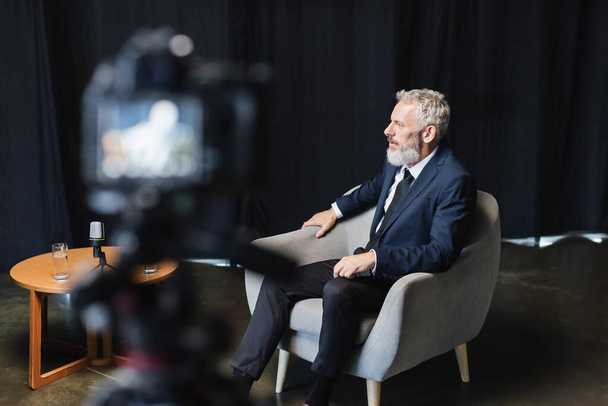 businessman in suit sitting in armchair during interview near blurred digital camera - Photo, Image